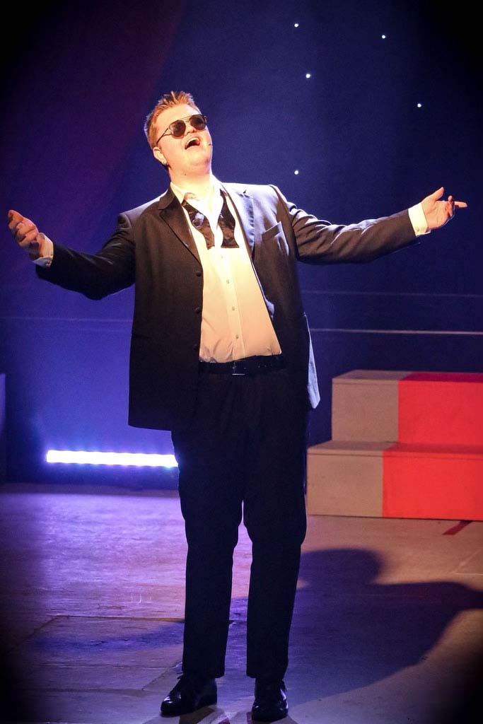 A performer in a dark suit and sunglasses sings with arms outstretched under blue stage light